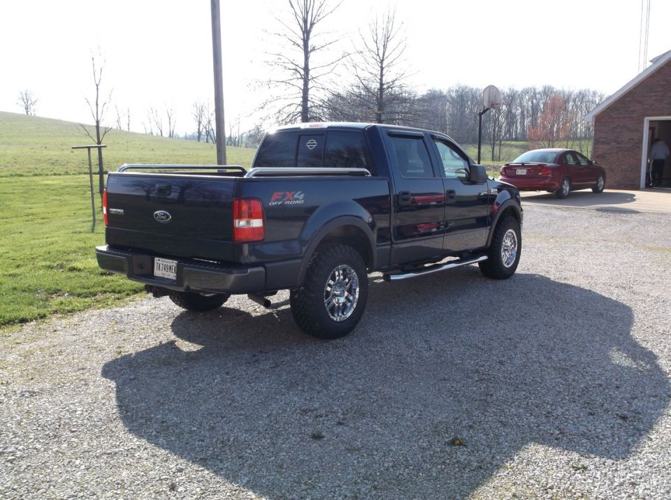 Dark blue Ford F-150 pickup truck with FX4 Off Road badging, parked on gravel. Red sedan in background
