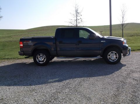 2005 Ford F-150 - Dark blue Ford F-150 pickup truck with aftermarket wheels and chrome side steps parked on a gravel lot.