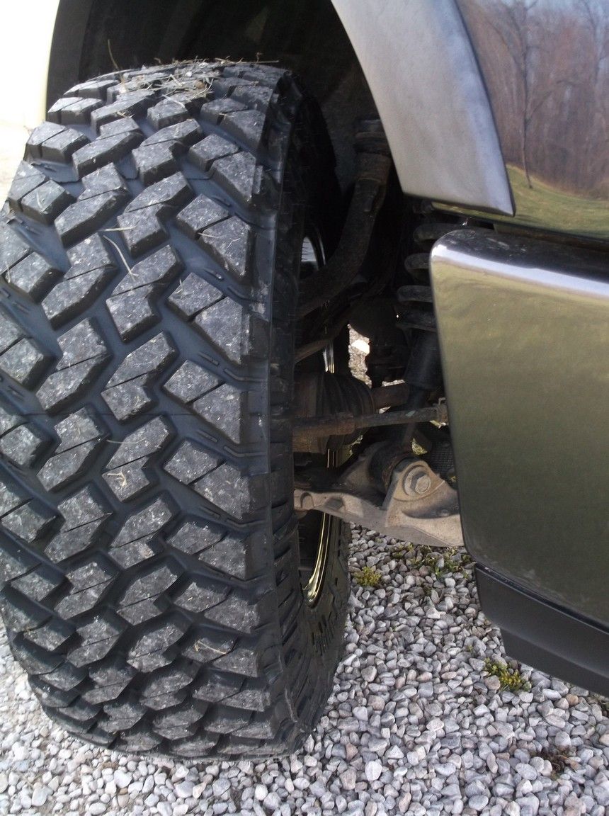 Close-up of a large, aggressive tread tire on a vehicle's front suspension. The tire is dark gray, and the suspension components are visible