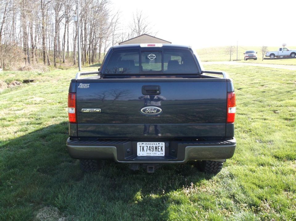 Rear view of dark blue Ford F-150 pickup truck with Indiana license plate TK 749 MEK. Another white pickup truck is visible in the background