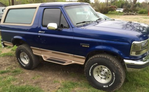 1996 Ford Bronco - This blue Ford Bronco features tan trim and running boards, parked outdoors on a grassy area.