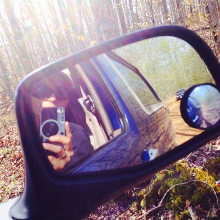 Person taking a selfie in a blue car's side mirror, another vehicle visible on a dirt road ahead