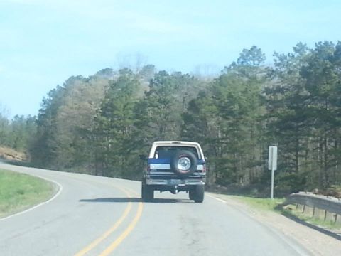 1996 Ford Bronco - Rear view of a blue and white Ford Bronco with a spare tire mounted on the back, traveling on a highway.