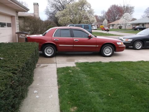 1999 Mercury Grand Marquis - A red sedan is parked in a driveway next to a hedge, with another vehicle visible behind it.