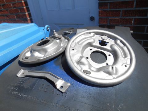 1972 Ford Bronco - Two brake backing plates and a metal bracket are laid out on a dark surface, with a blue trash bin visible.