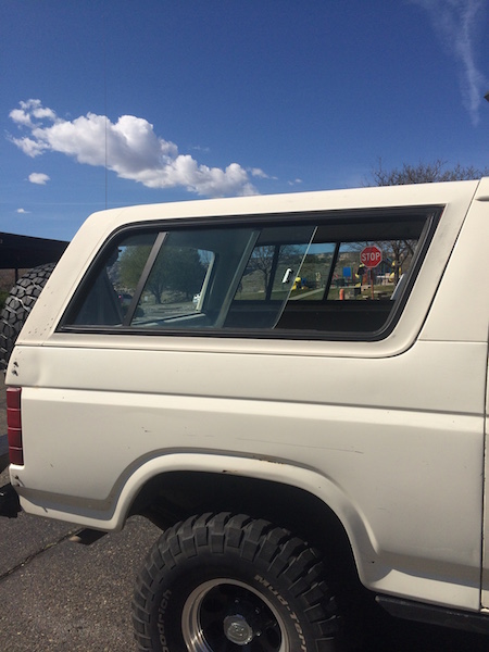 1981 Ford Bronco - The side view of a white Ford Bronco with a camper shell is shown against a blue sky with scattered clouds. The vehicle has large off-road…