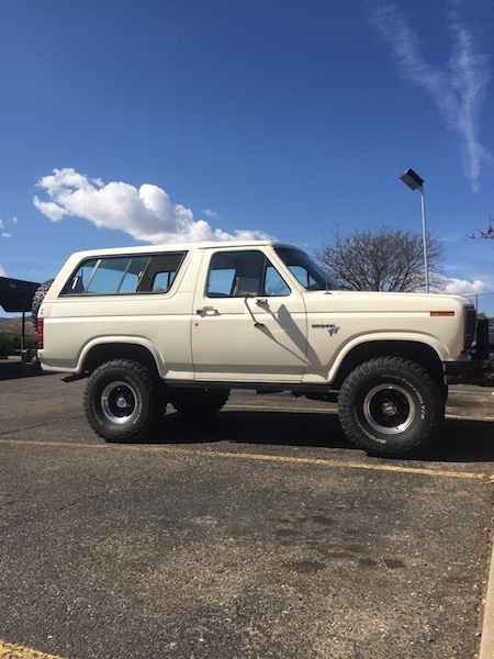 1981 Ford Bronco - A white Ford Bronco is parked in a lot under a bright blue sky with scattered clouds. The SUV has large off-road tires and is seen from the…