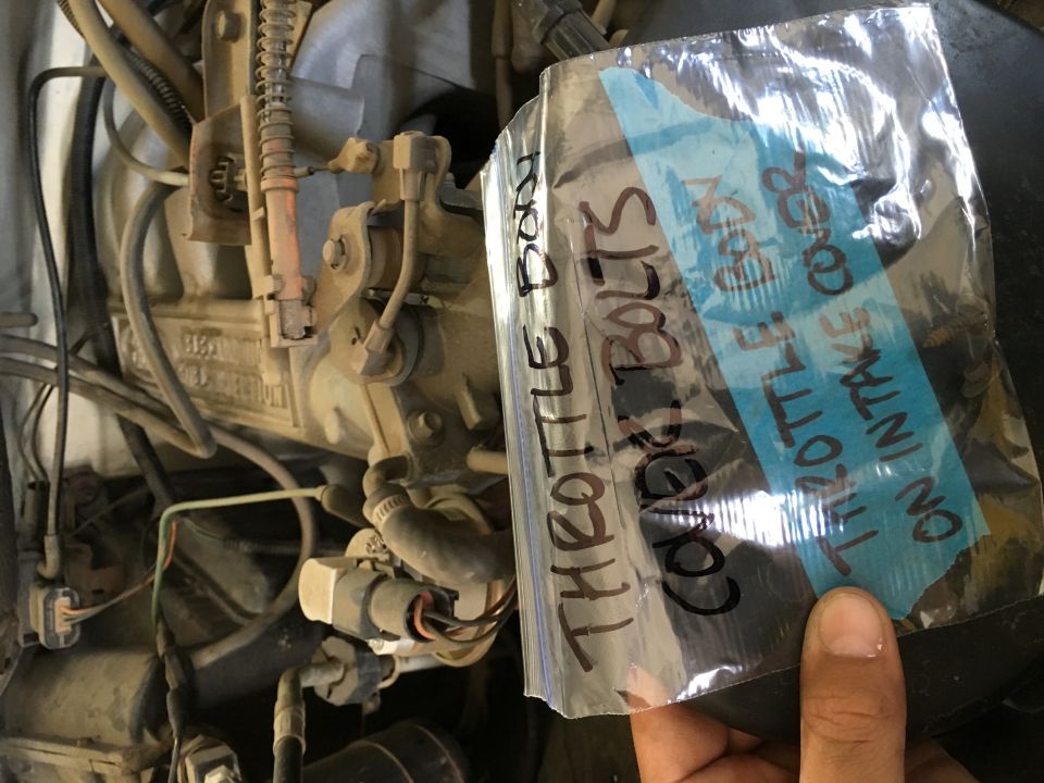Hand holding a plastic bag with text about throttle body cover bolts over a dirty engine.