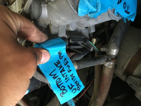 1987 Ford Bronco - A hand holds a piece of blue tape with handwritten text that reads "BOTTOM OF INTAKE BESIDE FUEL PM" over a section of an engine bay. The t…