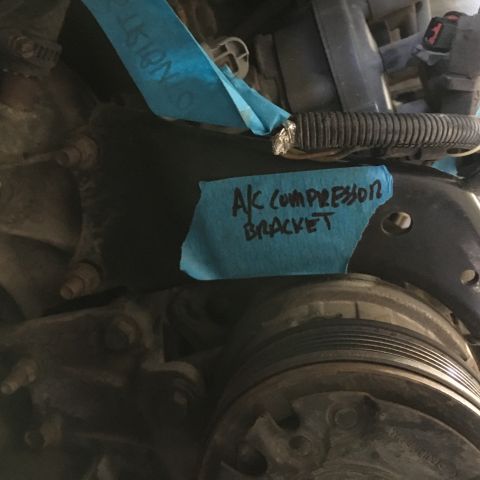 1987 Ford Bronco - A close-up view of an engine bay shows a black A/C compressor bracket labeled with blue tape. The bracket is secured by several bolts and i…