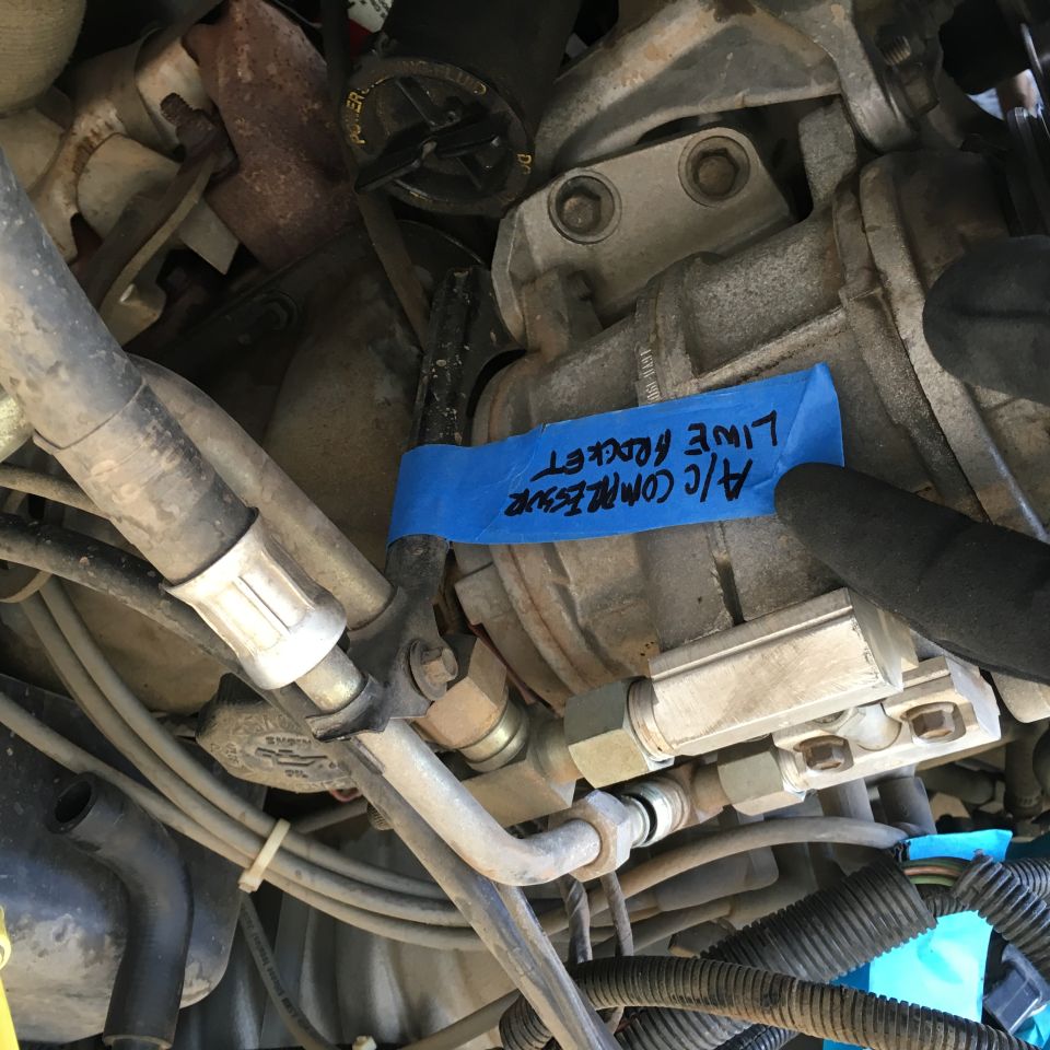 Close-up of a vehicle's A/C compressor with a blue tape label.