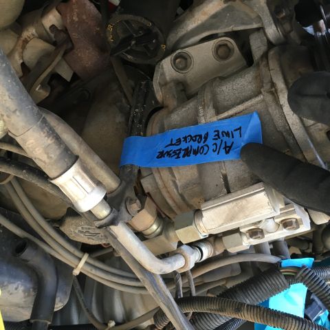 1987 Ford Bronco - A close-up view of an automotive engine bay shows an air conditioning compressor with a blue piece of tape labeled "A/C COMPRESSOR LINE HOC…