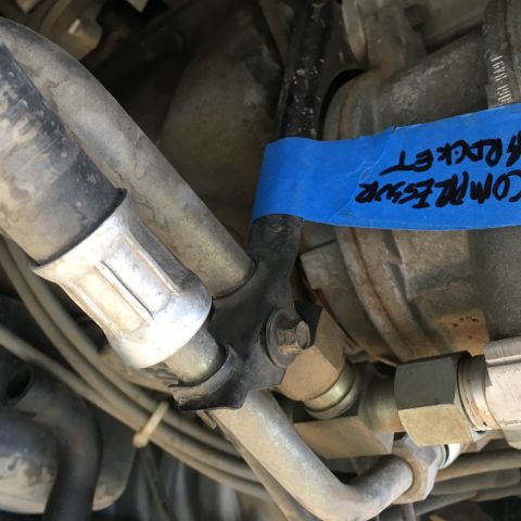 1987 Ford Bronco - A close-up view of automotive air conditioning lines connected to a compressor. Blue tape with handwritten text "COMPRESSOR ROCKET" is visi…