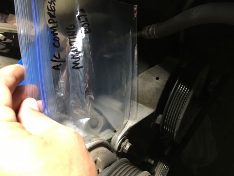 1987 Ford Bronco - A hand holds a plastic bag with handwritten text that reads "A/C compressor mounting bolts" over a portion of an engine bay. Visible engine…