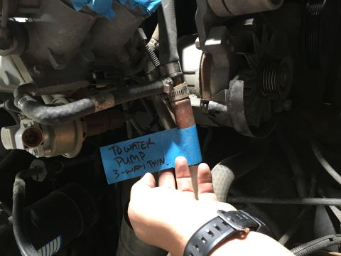 1987 Ford Bronco - A close-up view of an engine bay with a blue sticky note that reads "TO WATER PUMP 3-WAY THIN". A hand wearing a black watch is holding the…
