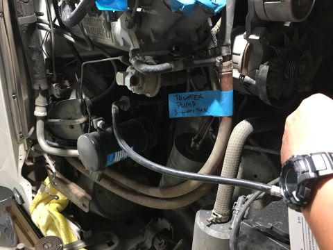 1987 Ford Bronco - A close-up view of an engine bay with a blue note attached to a hose that reads "TO WATER PUMP 3-WAY THIN". A hand wearing a watch is visib…