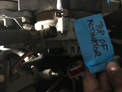 1987 Ford Bronco - A blue piece of tape with handwritten text reading "TOP OF ALTNATOR" is affixed to an alternator in a vehicle engine bay. A hand is holding…