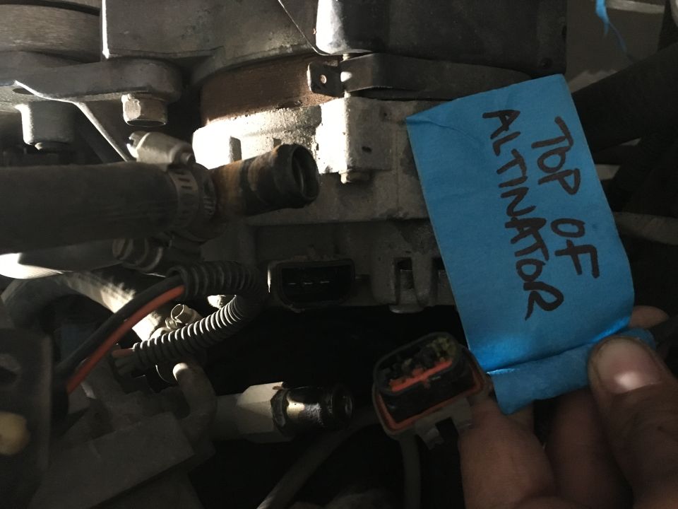 Close-up of a vehicle alternator with a blue tape label and a hand holding a connector.