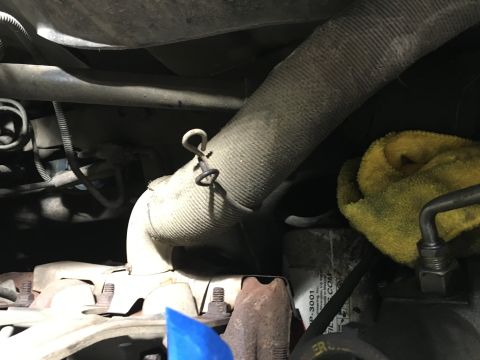 1987 Ford Bronco - A close-up view of a section of an engine bay, focusing on a heat-shielded exhaust pipe secured with a wire clip. A yellow microfiber cloth…