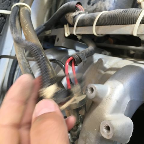 1987 Ford Bronco - A hand is shown disconnecting a wiring harness with red wires from a sensor on an engine. The engine bay is dusty and shows various hoses a…