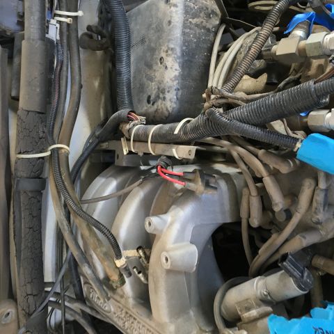 1987 Ford Bronco - A close-up view of a dusty engine bay shows a silver intake manifold with various hoses and wiring harnesses attached. Several electrical c…