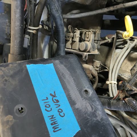 1987 Ford Bronco - A black plastic engine cover with blue tape and handwritten text reading "MAIN COIL COVER" is placed in the foreground, obscuring part of t…