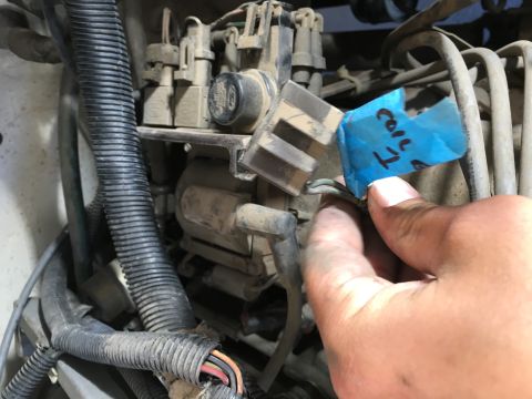 1987 Ford Bronco - A hand holds a piece of blue tape with writing on it, attached to a wiring harness near engine components. The tape is labeled 'Coil 1'.