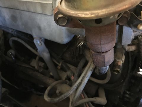 1987 Ford Bronco - A close-up view of engine components, including a carburetor or distributor with visible rust, and various hoses and wires.