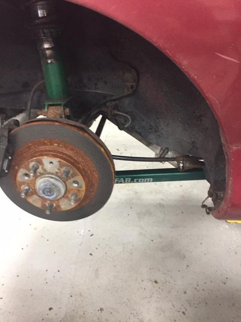 1995 Ford Probe - A close-up view of the front driver's side wheel well of a red vehicle, showing a rusty brake rotor, a green coilover suspension, and part…