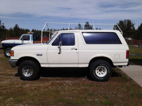 1990 Ford Bronco - A white 1990 Ford Bronco is parked on a grassy area next to a blue and white pickup truck. The Bronco has a roof rack and aftermarket wheel…