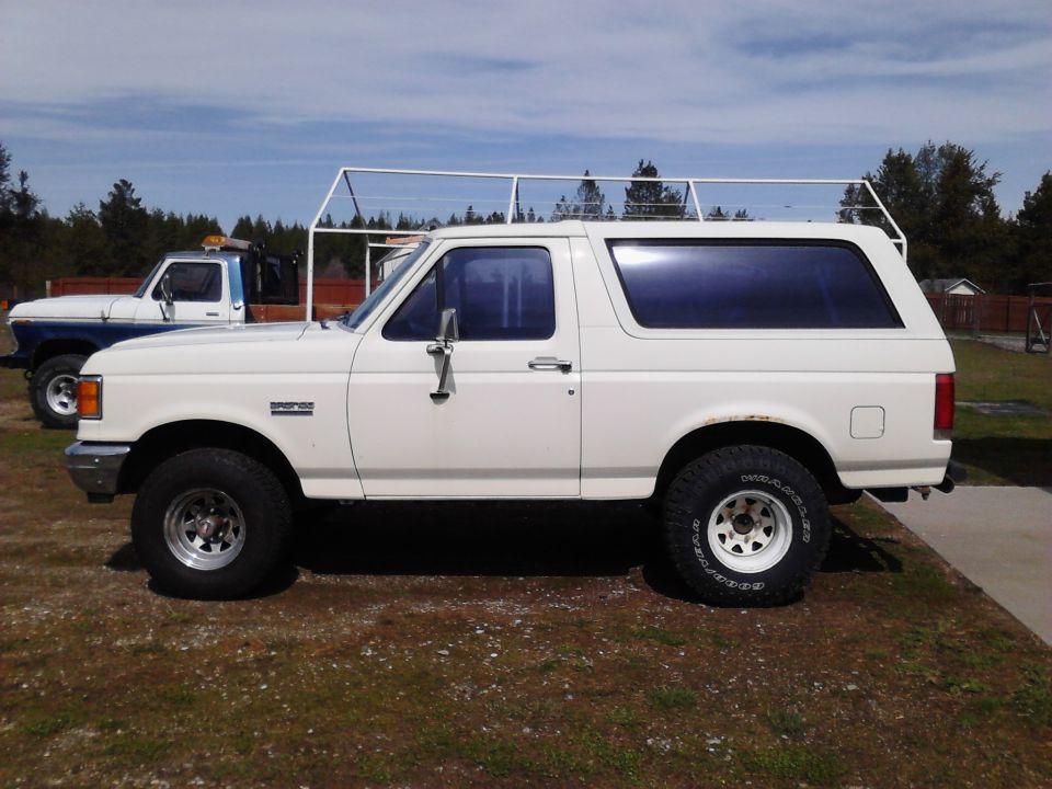 White 1990 Ford Bronco with roof rack and aftermarket wheels, parked next to a blue and white pickup truck.