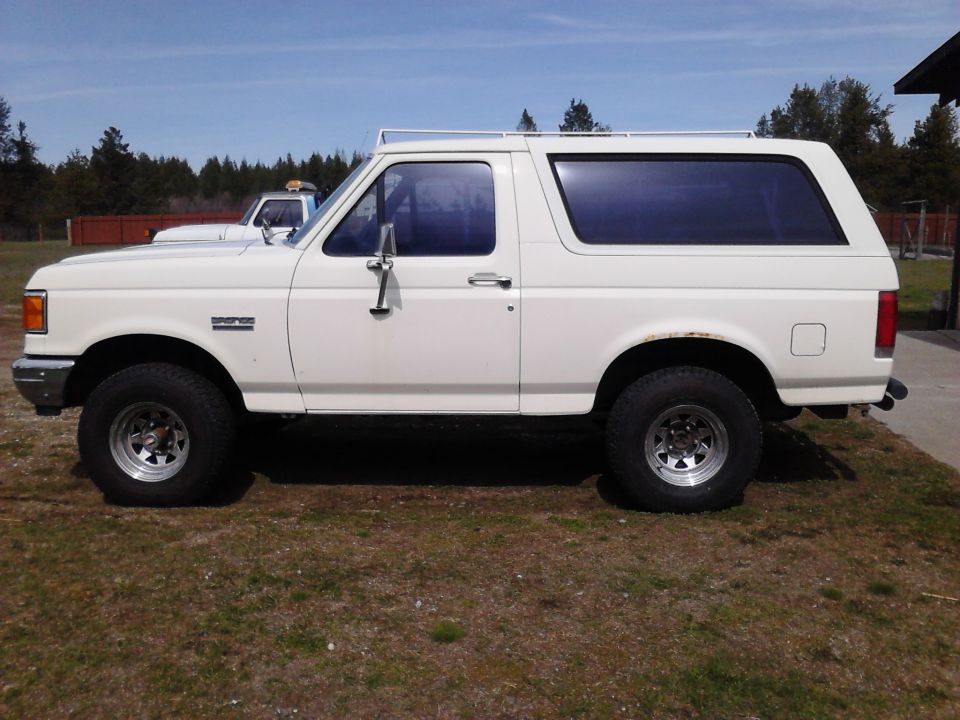 White 1990 Ford Bronco side view with chrome wheels and roof rack.