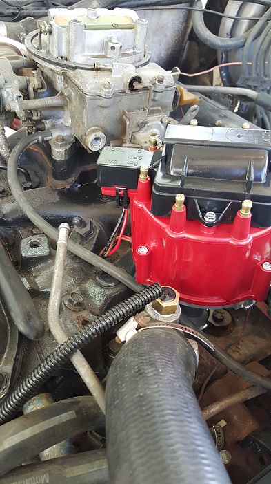 1978 Ford Bronco - A close-up view of a red ignition distributor cap and a carburetor on a vehicle engine. Wires and hoses are visible, along with part of a b…