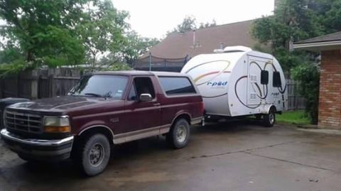 1996 Ford Bronco - A maroon 1990s Ford Bronco is parked on a paved surface, hitched to a white teardrop trailer. The trailer has a curved design with black an…