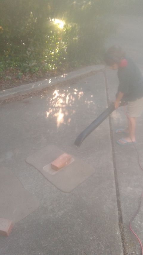 1996 Ford Bronco - A person wearing headphones is using a leaf blower on a driveway. There are car floor mats and a brick on the ground.