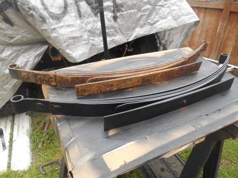 1972 Ford Bronco - Two sets of leaf springs are displayed on sawhorses outdoors. The top set is heavily rusted, while the bottom set is newly painted black.