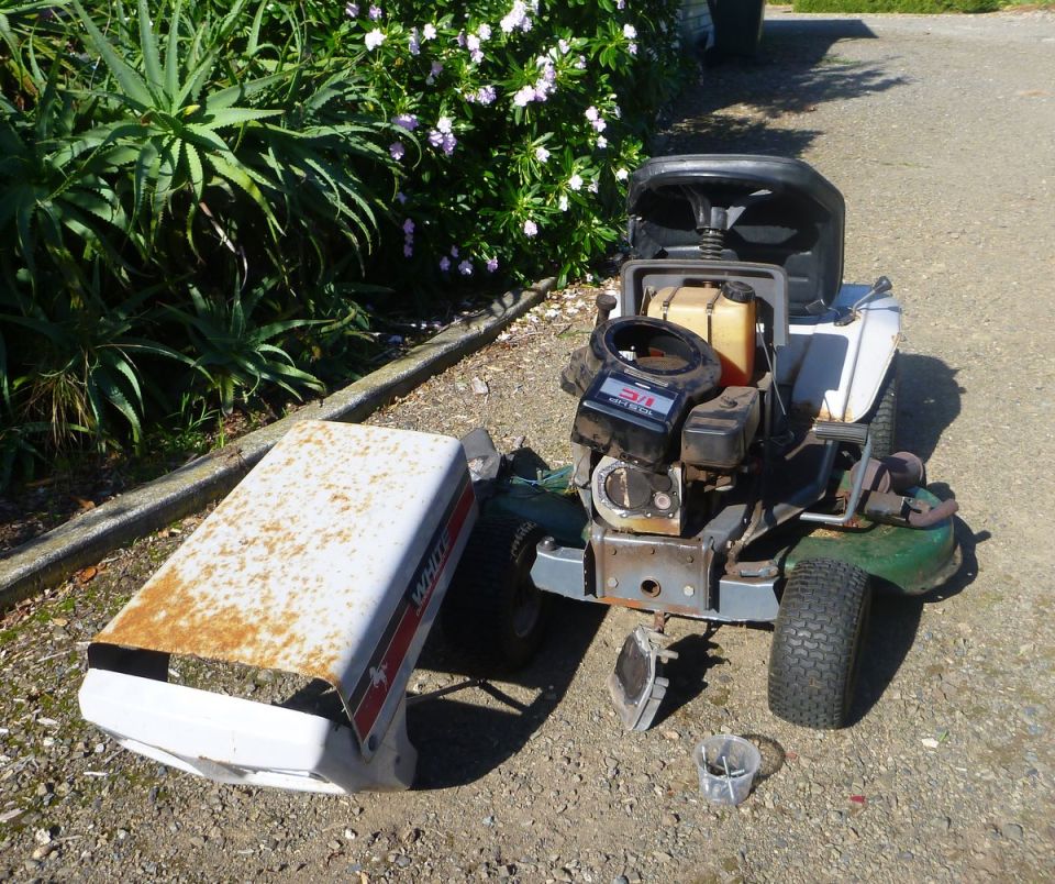 Riding lawn mower with hood removed, sitting on gravel next to some plants. Engine is exposed