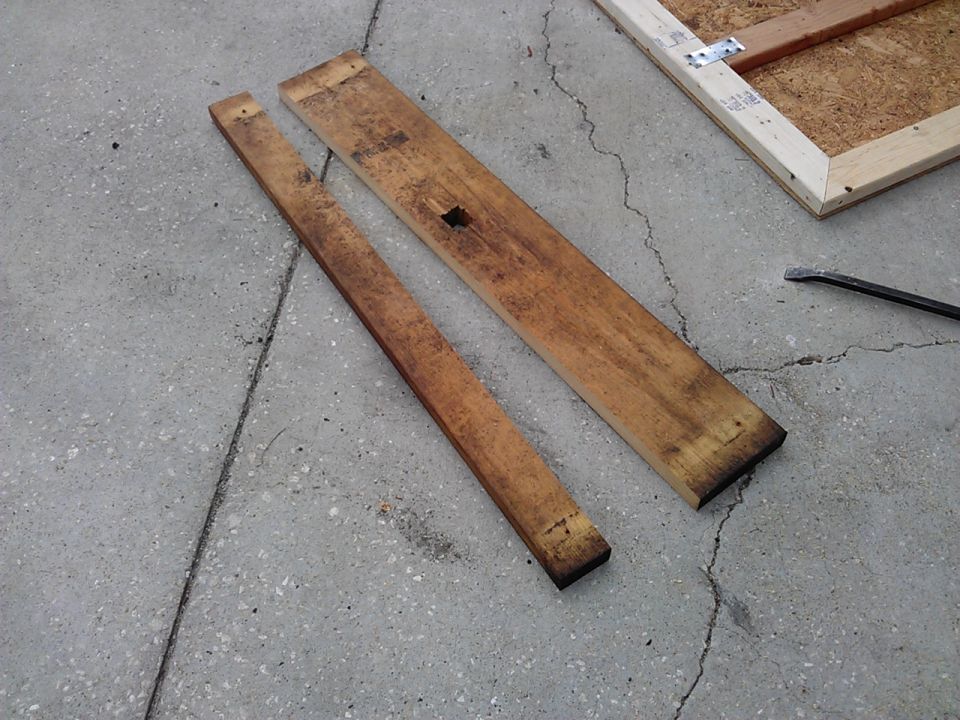 Two wooden planks lie on concrete next to a wooden frame with metal brackets. A pry bar is visible