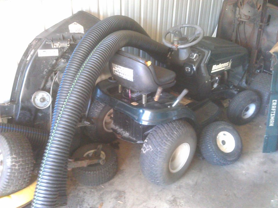 Dark green riding lawn mower with two large black hoses attached, parked indoors with a spare tire nearby.