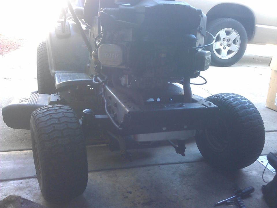 Rear view of a riding lawn mower with a large engine and one rear tire in focus. Car in background.