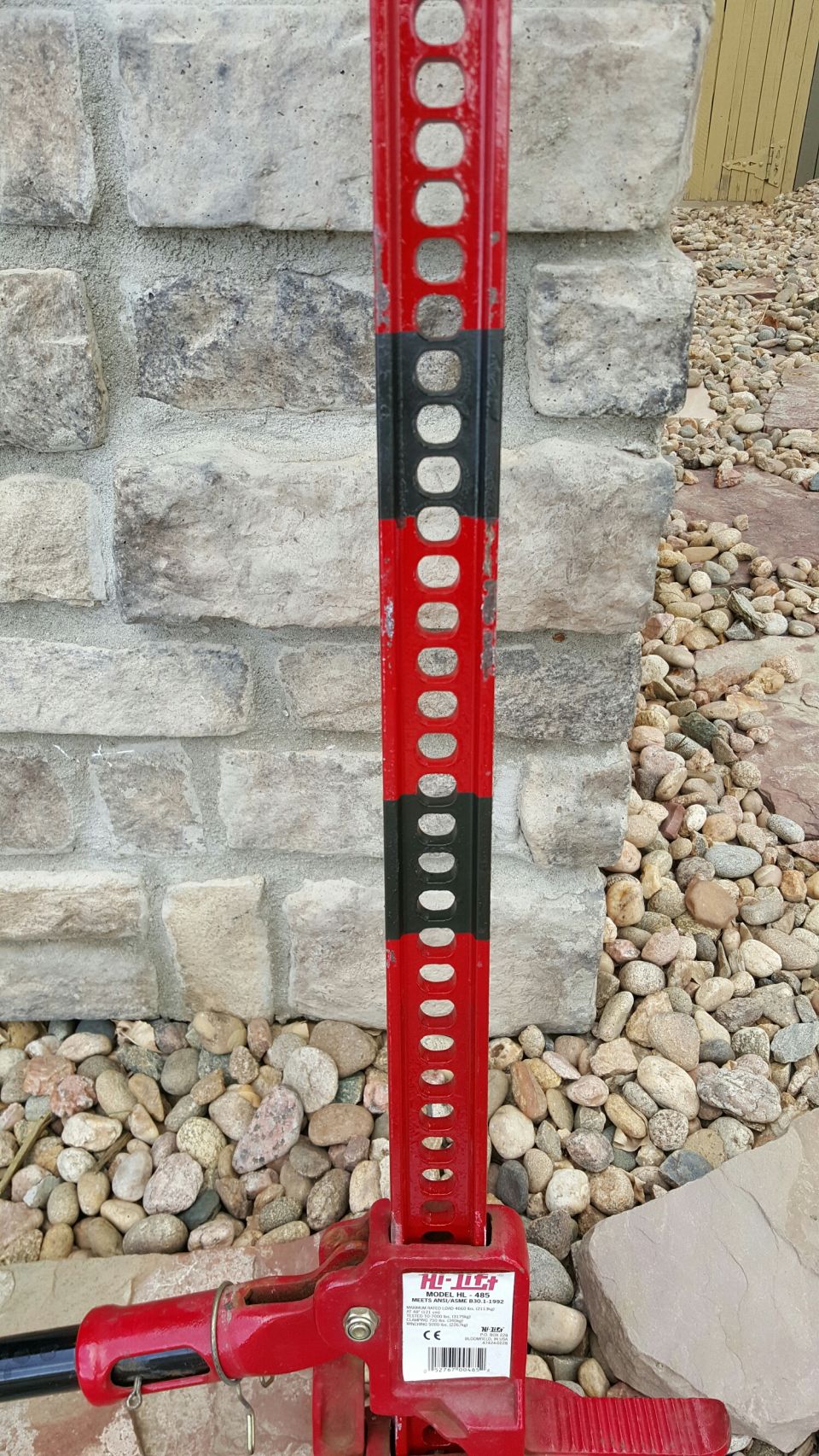 Red and black Hi-Lift farm jack leaning against a stone wall.
