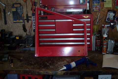 1990 Ford Bronco - A red toolbox with a red door frame leaning against it sits on a cluttered workbench. A tube of caulk and a caulking gun are in the foregro…