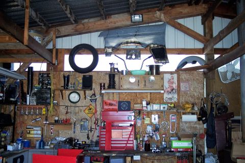 1990 Ford Bronco - A cluttered garage workshop is filled with tools, car parts, and various items hanging on the walls and shelves. A large tire hangs from th…