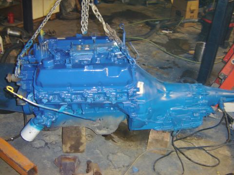 1971 Ford Ranchero - A bright blue Ford V8 engine and automatic transmission assembly is suspended by chains in a workshop. The engine is supported by wooden bl…