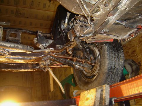 1971 Ford Ranchero - Underside view of a vehicle's front suspension and wheel, showing rusted metal components and a tire resting on wooden blocks.