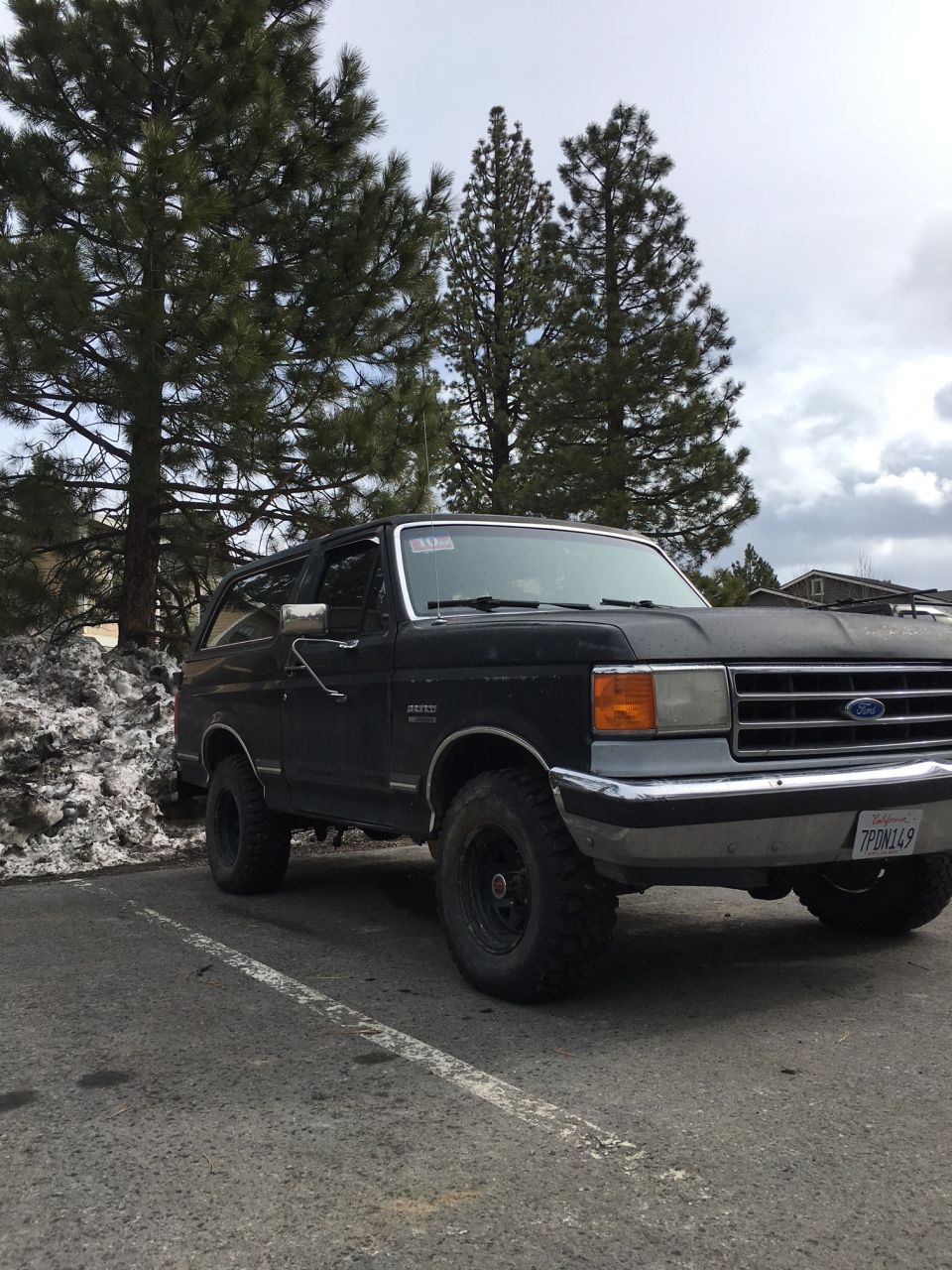 Black 1988 Ford Bronco parked in a lot with snow and pine trees.