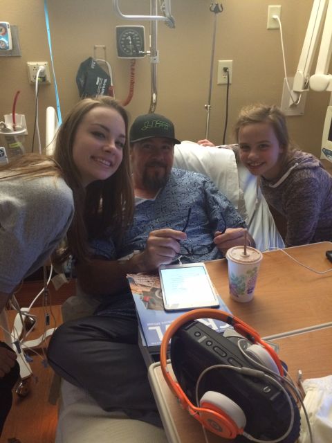 1985 Jeep CJ7 - A man in a hospital gown is sitting in a hospital bed, with two young women smiling beside him. A tablet, a cup, and headphones are on a be…