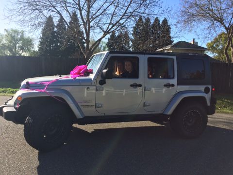1985 Jeep CJ7 - A silver Jeep Wrangler Unlimited is decorated with a large pink bow and ribbon on the hood. Two young women are visible inside the vehicle.