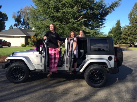 1985 Jeep CJ7 - Three young women stand around a silver Jeep Wrangler with the doors open. The Jeep is parked on a paved driveway in front of a suburban ho…