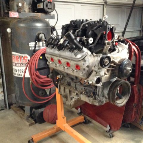 1985 Jeep CJ7 - A V8 engine is mounted on an orange engine stand in a garage setting, next to a large air compressor and a red air hose.
